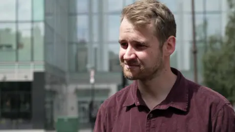 A man in a burgundy shirt standing outdoors in front of a contemporary glass-fronted building.