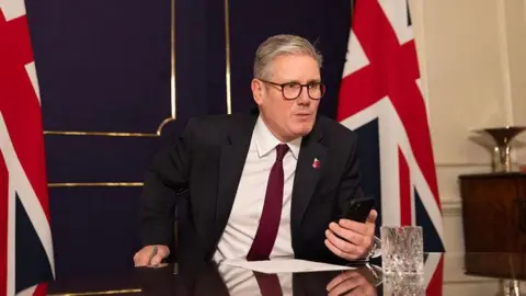 Downing Street Sir Keir Starmer holsd his mobile phone in front of him as he speaks to Donald Trump from his office in No 10 Downing Street. There are two union jack flags behind him.