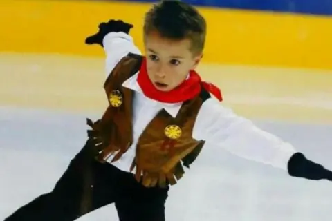 Karen Brennan A young Edward stretches out in a star shape while on the ice. He is wearing a cowboy costume with a red neckerchief. There is a look of deep concentration on his dace.