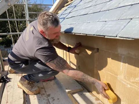 a tattooed man wearing a t-shirt sponges down sandstone while standing on scaffolding at the church. You can faintly see an engraving on one fo the stones saying High Toynton 2025