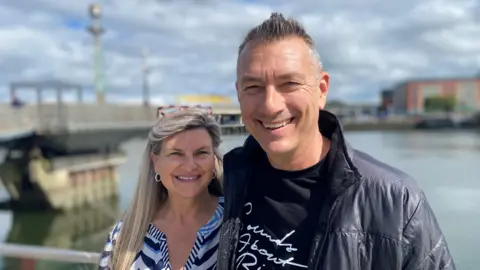 On the left, a long-haired blonde woman in a striped blouse. On the right, a tall, dark-haired man in a black puffer coat and a black t-shirt with white writing.  