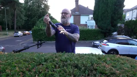 A man wearing a purple shirt holding up a ring he has just found on top of a bush.