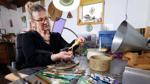 A woman with blonde hair pulled back off her face and glasses in a dark top with a large bead necklace works over a blowtorch in a studio littered with items to help her artwork