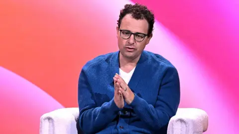 Getty Images Dario Amodei sitting on-stage at a conference, wearing a blue cardigan and white t-shirt. He is holding his hands clasped in front of him as he speaks.