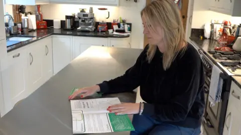 A woman wearing a black jumper sits at a kitchen island and reads a white and green booklet