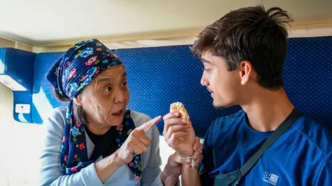 BBC/Studio Lambert An elderly woman holds Kush's hand as she tells him to eat a food item while they on a train in Kazakhstan.