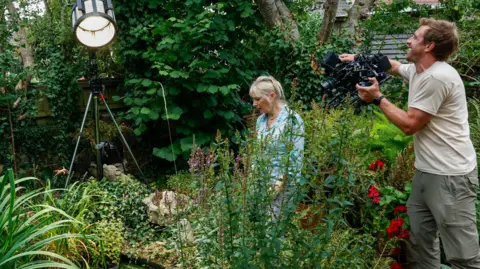 Louise Vergette wearing a blue and white blouse, with her blonde hair tied up. She is walking through her garden, which is full of different types of trees, flowers and plants. 