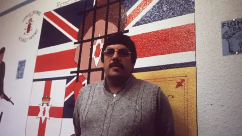 Pacemaker A man in a grey woollen sweater and dark sunglasses is stood in front of a mural consisting of loyalist insignias and flags across a British Union Jack.