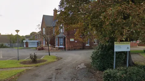 A red brick building away from a main road. It has a big car park outside. A sign says: "North Lincolnshire Council, The Hollies, The Cygnets.
