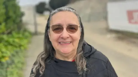 Jill smiling at the camera. The background is blurry, but an outdoor scene is visible behind her. She has shoulder length grey hair and is wearing glasses.