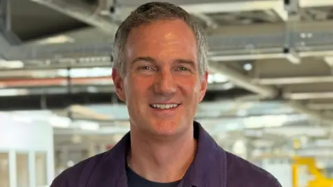 Peter Kyle stand with the Rolls-Royce manufacturing floor behind him. He is smiling and has grey hair and blue eyes. He wears a black t-shirt beneath a purple jacket. 