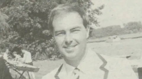 Henley Standard A black and white photo of two men dressed in blazers with striped ties grinning at the camera. Behind them is a river with several people working on rowing boats. 