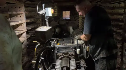 A worker is wearing a dark T-shirt and gloves operating a large piece of machinery inside a dimly lit industrial workshop. Wooden racks stacked with rectangular tiles line both sides of the narrow room, creating a corridor-like space. A single overhead work light illuminates the machine and the worker’s hands, while additional equipment is visible in the background. The scene highlights a traditional manufacturing process in a busy, enclosed workspace.