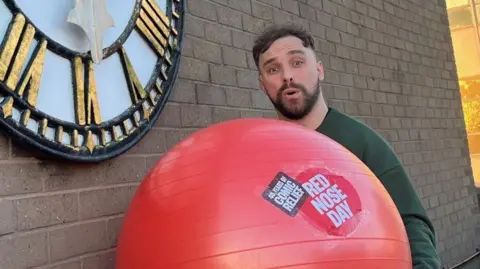 Lee Blakeman, on the roof of the BBC Radio Stoke building, holding a large red medicine ball with Red Nose Day branding on it. He has short dark hair and a dark beard and stands next to a wall with a clock on it.