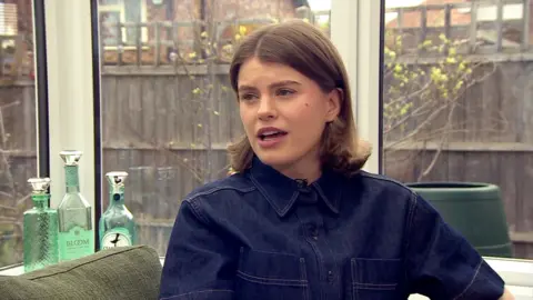 A white women with brown hair and a demin top sits talking to someone in a conservatory.