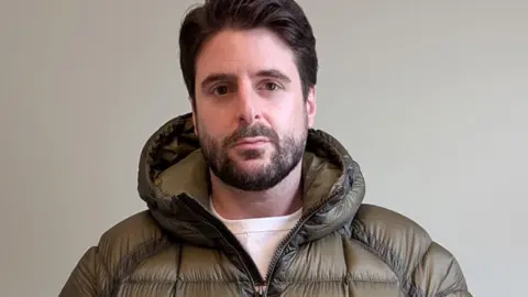 Alex Grossomanides has brown hair, brown eyes and a beard. He is wearing an olive green puffer coat and looking at the camera while standing in front of a white wall indoors.