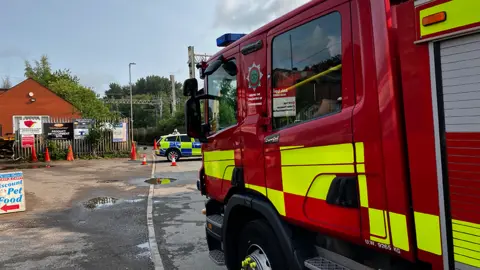 The side of a fire engine fills the right hand side of the image. A police car is in the middle of the photo in the distance, near to cones and signs on railings. There is a small brick building to the back left of the image behind a high railing. 