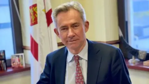 A man wearing a suit and tie sitting at a desk with a Jersey flag behind him and windows.