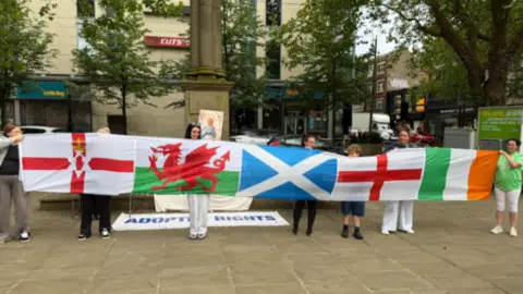 People at a vigil for murdered baby Preston Davey in Preston's Flag Market holding up flags. People are starting to arrive at the vigil to remember the 13-month-old. An image of Preston can be seen in the background and a sign saying 'Adoptee Rights'. 