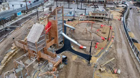 Esh Construction/Chapman Brown Photography A construction site with a large timber play structure in the centre. The play towers have large steel slides attached. Orange rope netting bridges can be seen as well as wooden walk ways. The picture was taken on a wet day and muddy puddles can be seen beside the construction work. 