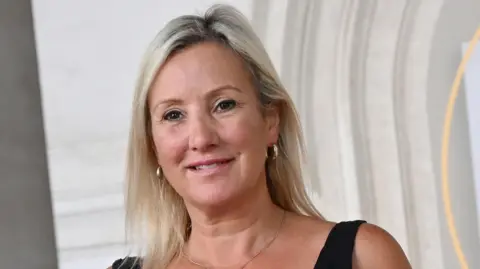 Getty Images Caroline Dinenage MP. She has blonde hair and is wearing a black top with shoulder straps. She has a gold hoop earring in each ear. 