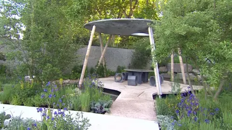 At the garden's centre sits a solar-powered shell-shaped canopy with a metal round roof and large struts holding it up. It is surrounded by greenery