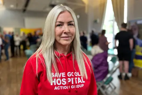 BBC/Anne-Marie Tasker A woman with long blonde hair and a red top, with "Save Goole Hospital Action Group" written on it. There are people standing in a room behind her.