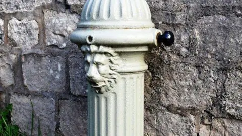 Historic England A green iron water fountain with a lions head. It is standing against a dark stone wall