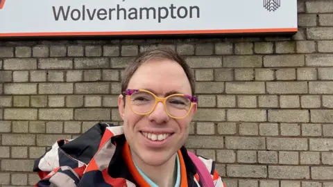 Alex Manners A man in a orange, white, beige and black jacket, with purple and yellow glasses, stood in front of a brick wall with a Wolverhampton sign.