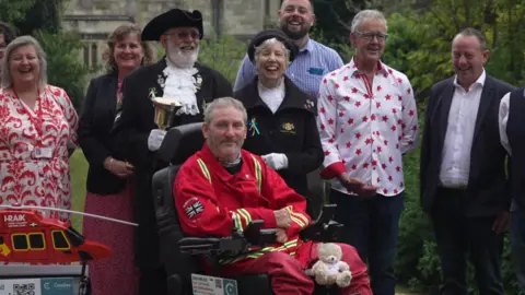 A photo which shows Steven Webb in the centre in an electric wheelchair. He is wearing a red Cornwall Air Ambulance outfit. There is also a small teddy bear on his lap. There are people behind him, including a town crier holding a bell and wearing a hat.