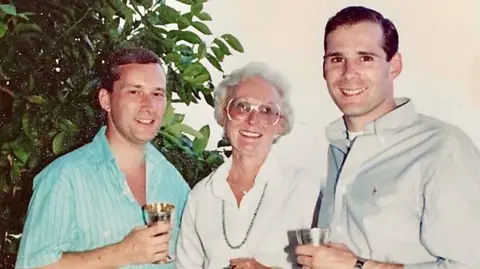 Cressman Family Pictured from left to right is a man wearing a blue shirt, a woman wearing a white shirt and a man wearing a light blue shirt. All three are holding drinks and looking towards the camera.