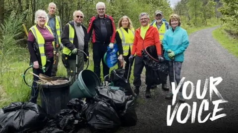 BBC A group of eight volunteers wearing hi-vis vests stand behind a pile of rubbish bags piled up beside a n overflowing bin amongst the green landscape around Loch Lomond.