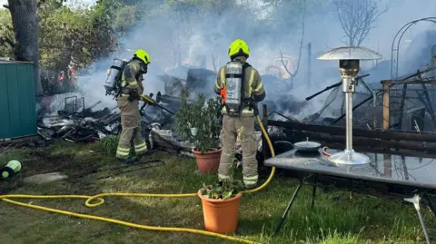 Essex Fire and Rescue Service Firefighters standing with their back to the camera with a hose in a grassy garden area with potted plants and a glass table. In front of the firefighters there are the remains of a burnt out home. 