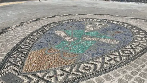 Tom Jackson/BBC A mosaic depicting the 9th Century Saint Neot. It is inlaid into a pavement and shows a stylised male in white, grey, blue and green stones and tiles. A grey stone plaque at the bottom has the words St Neot on it