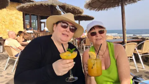 Delia Lodge Rebecca is on the left and Delia is on the right. The pair are pictured on a beach drinking a cocktail each. 