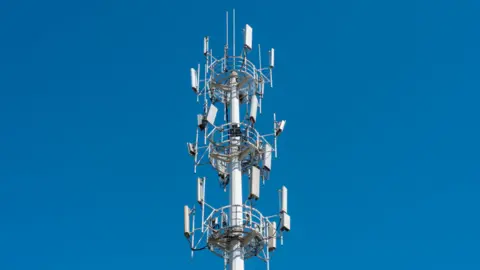 A telecommunications tower fitted with multiple antenna panels rises against a clear, bright blue sky. The structure is tall and metallic, with several circular platforms and railings positioned at different heights. Rectangular antenna units extend outward in multiple directions around the tower, along with smaller antenna rods and equipment mounted near the top. The image focuses tightly on the upper portion of the tower, with no surrounding buildings or landscape visible.