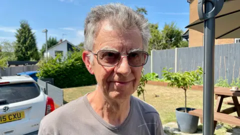 A man with grey hair and glasses wearing a grey t-shirt stands in a sunny back garden. Behind him are a parked car, a wooden picnic bench and raised garden beds. He is looking directly at the camera.
