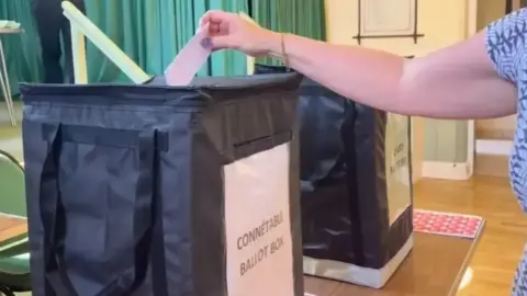 BBC A person putting their vote into a black electoral box at a village hall. A green curtain behind. The woman is wearing a blue dress.