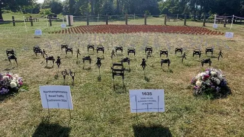 Black silhouettes representing people, cars and bicycles planted into grass. There are notices planted in front showing 1635 - the number of people killed or injured on the county's roads in 2024. There are bouquets of flowers on the left and right. There are grey dove silhouettes behind and small red county flags behind those.