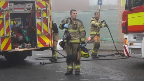 A firefighter dressed in scorched yellow clothing holds a yellow helmet and mask. In the background, another firefighter moves a hose.