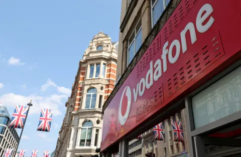 Reuters A street scene looking up at shop with a red sign with Vodafone printed in white letters alongside a red and white, circular logo. In the background, a blend of vintage brick and modern glass buildings can be seen, along with Union flags hanging across the street under a blue sky.