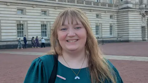 A woman standing outside Buckingham Palace. She is smiling. She has long light brown hair with a fringe. She is wearing a green dress with a necklace around her neck.