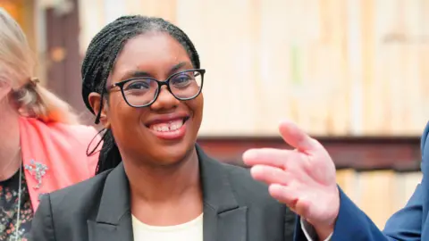 A woman wearing a grey blazer and a white top and smiling at the camera.
She is wearing black frame glasses 