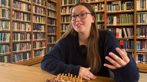 A white teenage girl with long light brown hair, wearing dark-rimmed glasses and a navy jumper, sitting at a desk with a chess board on it, in front of two large bookcases