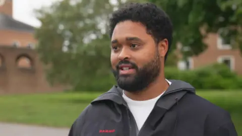 Solomon Elliot looking to the left and smiling. He is wearing a white top and a black jacket.