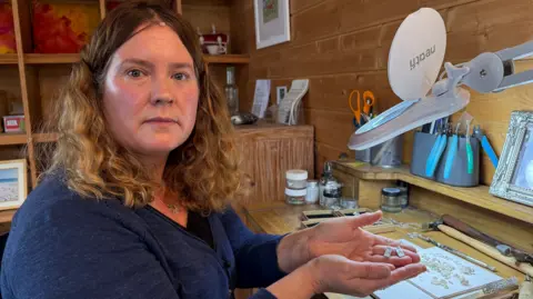 A woman with brown curly hair wearing a blue jumper. She is sitting in a wooden workshop surrounded by various tools. She is holding a pair of silver earrings which are rectangular in shape.