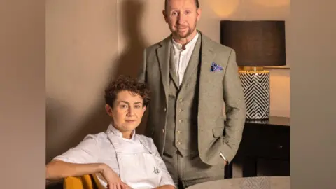 Hansom Restaurant A woman wearing a white chef shirt and a white apron sits on a wooden chair. She has brown hair, and has one arm resting against the back of the chair as she turns toward the camera. Standing by the chair is a man wearing a sage green three-piece suit over a white shirt. He has his hands in his pockets, and a pocket square in his top pocket.