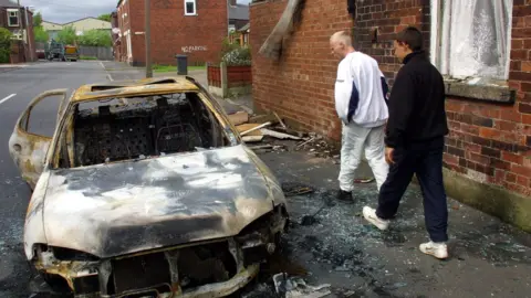 Two young people pass by a burnt out car wreck in the northern English town of Oldham 29 May 2001.
