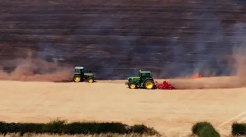 Farmers in green tractors create firebreaks to stop the spread of flames in a field.
