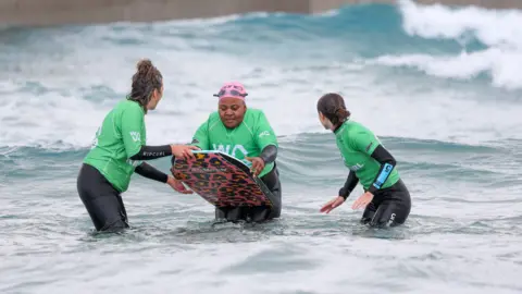 The Wave A participant getting ready to catch a wave on her bodyboard, joined on either side by two volunteers who are gearing up to help push her into the wave. The participant is wearing a black wetsuit, green rash vest and pink swimming cap with goggles on her head. Behind them there are two small waves about to break.